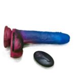 Dildo cósmico de silicone azul e rosa deitado lateralmente ao lado de seu controle remoto sem fio oval e preto.