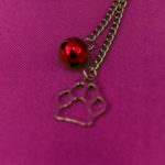 Detalhe em macro focando na extremidade da corrente de um acessório BDSM pet play, mostrando nitidamente um guizo metálico vermelho brilhante e um pingente prateado em formato de patinha de animal, sobre fundo verde texturizado.