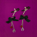 Par de prendedores de mamilo ajustáveis de metal prateado para fetiche pet play, com pontas emborrachadas pretas para proteção. Decorados com laço de cetim preto e correntes metálicas pendentes segurando um guizo vermelho e um pingente de patinha vazada, dispostos sobre um fundo verde vibrante.